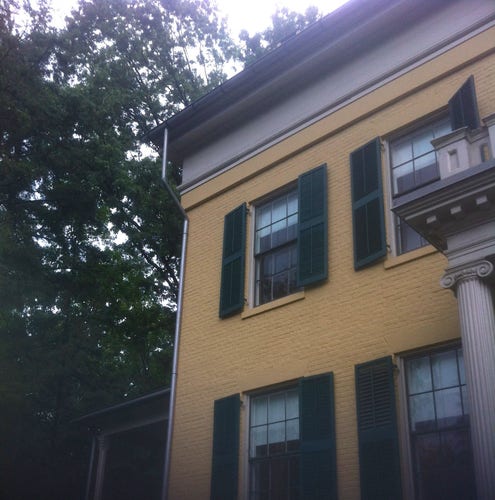 Yellow brick, two-story house with ordinary windows framed by dark green shutters. On the right, the left side of a columned portico at the front entrance.
