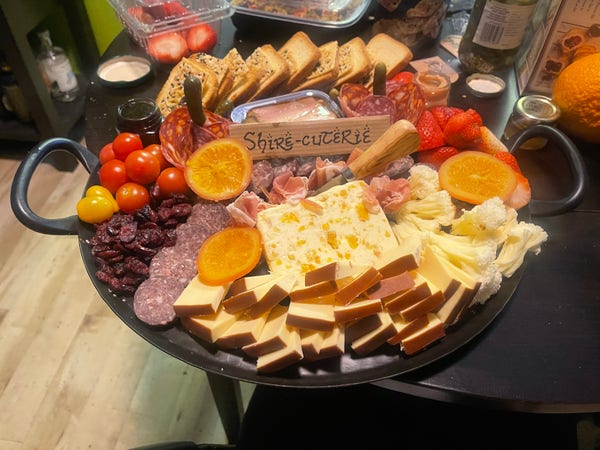 Giant charcuterie platter filled with cheeses, meats, fruits, crackers, & jams. A sign in the middle says shire-cuterie in a lord of the rings style typeface. 