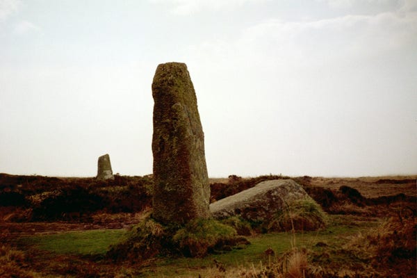 Colour photo of the Nine Maidens Stone Circle at Boskednan in West Cornwall. An upright stone stands in the foreground with a fallen stone beside it. In the distance is another of about the same size. The sky is grey.