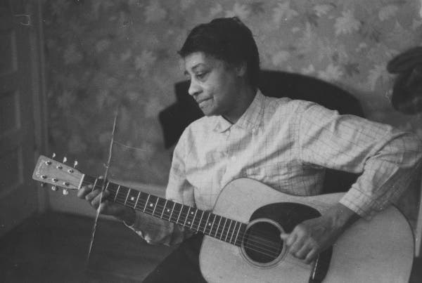 Elizabeth Cotten, fotografiada por John Cohen en 1960. Es una mujer afroamericana mayor, vestida con una blusa abotanada hasta el cuello y toca una guitarra mientras mira hacia el mástil dándonos su perfil. Puede verse que es zurda y la guitarra simplemente está girada. Smithsonian Folkways Recording.