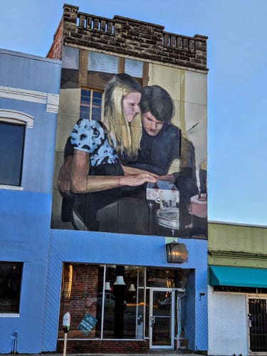 Above a small shop on the first floor of an old, tall brick building a mural of a young couple seated at a table, opening a small wrapped package together.