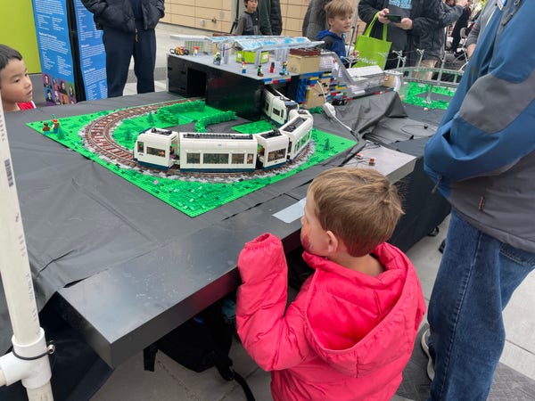 A child in a red jacket looks intently at a LEGO model train display with onlookers around and information boards in the background.