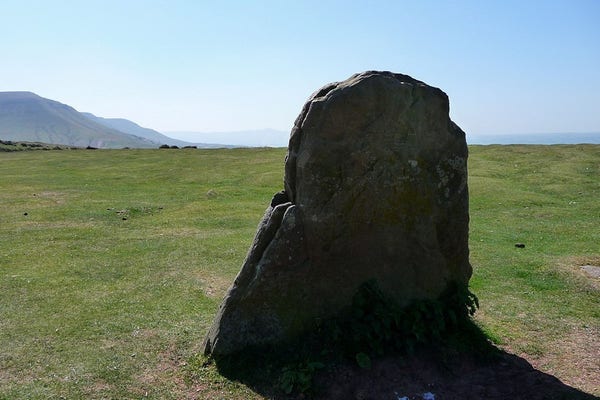 A short standing stone on a grassy plateau. The stone is a slab, thin on one face and broad on the other. It tapers from the broad base to a rounded top. The escarpment of a mountain range can be seen behind to the left, blue and hazy. The sky is clear blue and it's very warm.