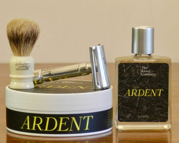 A tiny silvertip badger shaving brush bearing the signature of Alexander Simpson and an aluminum slant razor with a ribbed handle sit atop a tub of shaving soap whose black label has "Ardent" in large yellow letters. Next to it in a small rectangular glass bottom with a cap the same color as the razor. The bottle's label is black with "Ardent" in yellow, and also has in white letters at the top "The Razor Company."