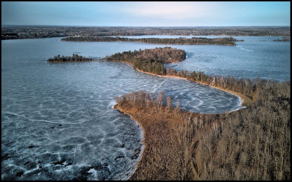 Aerial view of a frozen lake with several small islands, narrow penninsulas, and small bays. The vegitation is all very brown, except for some scattered evergreen trees. Almost no snow on the ground.