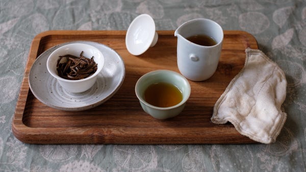 wooden tea tray with small porcelain gaiwan showing black tea leaves, in a ceramic cup, pitcher, green porcelain cup