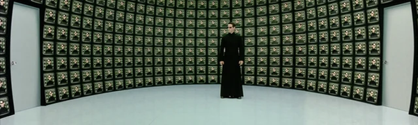Wideshot of Neo. Standing in front of a wall  of screens showing Neo in closeup.