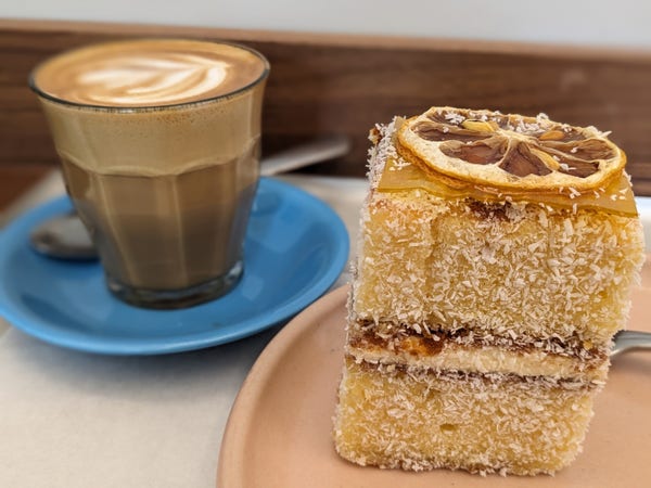 Colour photograph of a square piece of sponge cake, with a jam and buttercream filling, coated in coconut with a slice of dried orange on to, accompanied by a latte.