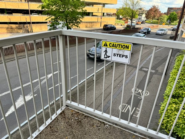 A brightly coloured warning sign, mounted on the inner railing of a pedestrian bridge across a busy road, says “Caution: Steps”