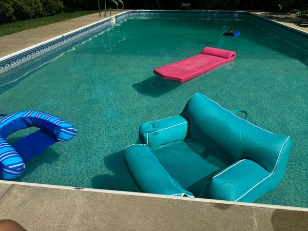 Three pool floaties bob in a large private pool. One is pink and made to be laid on, the others are blue and arm-chair-esque 