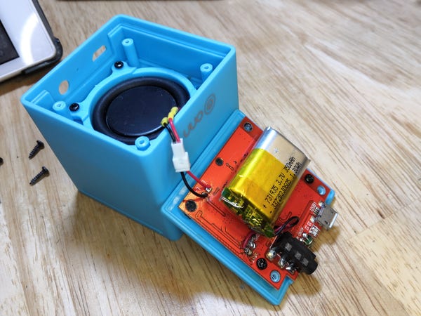 A light blue cube shaped Bluetooth speaker with the panel opened to reveal red PCB. The lithium battery has been replaced with a rectangular one from a disposable vape device. A JST plug has also been added to the speaker connection wires to make future disassembly easier