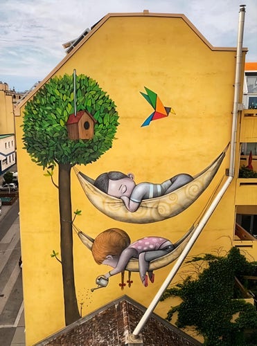 Streetartwall. A mural of two children sleeping in hammocks on a colorful tree was sprayed/painted on the upper part of a four-story house. The mural is painted from the second floor up, as a roof covers the lower part of the wall. The background is a cheerful yellow. On the right side is a green tree with a birdhouse painted in a naive style. A colorful, folded paper bird flies in the middle of the picture. Two painted hammocks are stretched out below it. In the upper one, a small dark-haired boy lies sleeping. In the lower one, a brown-haired girl is watering the tree with a watering can. Wonderful.
(On the wall is a long, light-colored, kinked rain gutter that leads down to the roof below. It has been integrated into the mural. It serves as a "support" for the two painted hammocks)