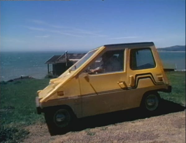 tiny, yellow, trapezoidal 2 seat car on a seaside cliff 