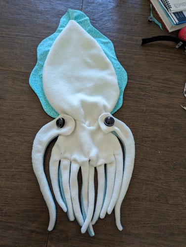 A partially sewn shell of a white fleece cuttlefish plush animal.wirh a large blue fin around its body. The cuttlefish has eight small tentacles and two longer arms hanging down from its body.