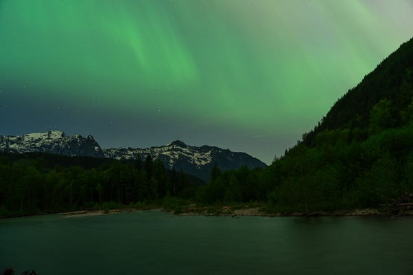 A photo of a landscape at night with snow covered mountains and a river in the foreground. The sky is ablaze with the aurora borealis with beautiful greens and yellows