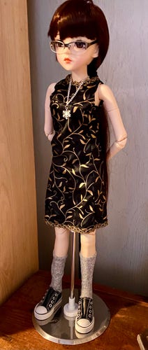 A fashion doll with brown hair wearing glasses, a black dress with golden patterns, a necklace, gray socks, and black sneakers, standing on a stand.