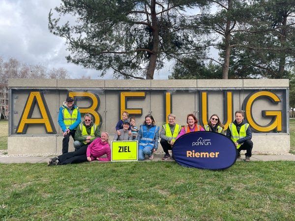 Photo of nine volunteers in yellow jackets squatting and lying in front of the Abflug sign in yellow letters. The "Ziel" and "Riemer" parkrun signs are also on display.