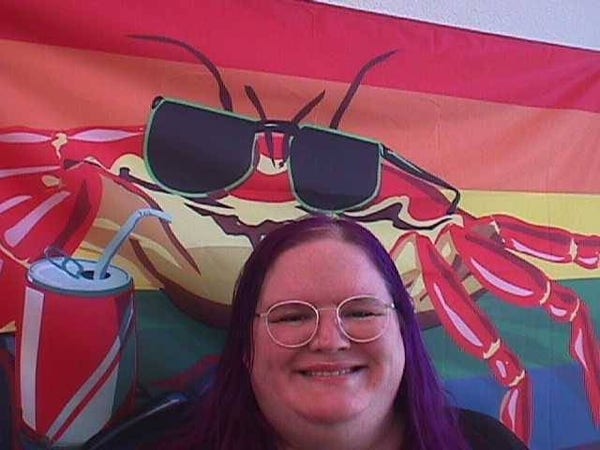 A purple-haired person with large round glasses smiling in front of a crab-on-a-pride-flag flag. 