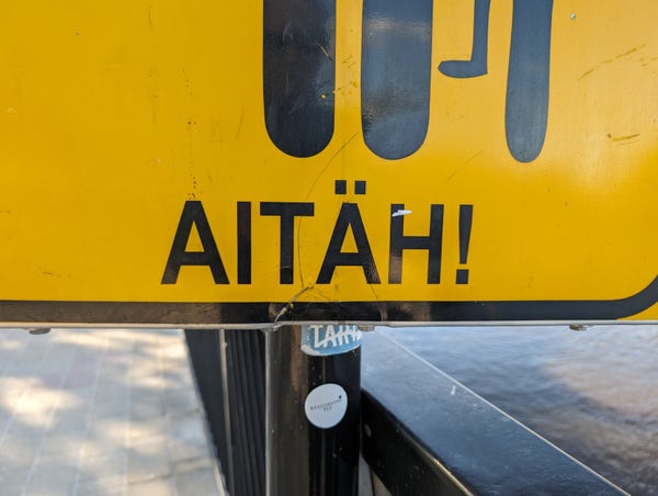 The bottom edge of a yellow cyclists dismount sign on the riverbank in Tartu, Estonia. It reads AITAH