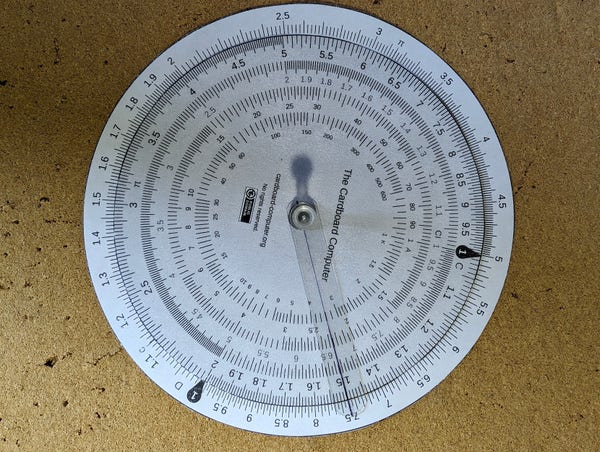 A paper circular slide rule pinned to a corkboard. The plastic cursor is over 1.5 on the C scale lined up with 7.5 on the D scale.