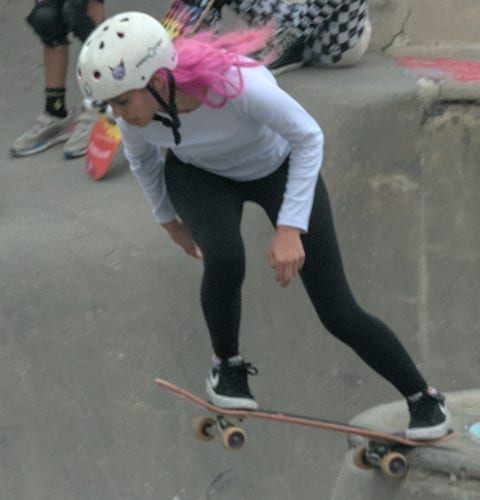 A young woman with bright pink hair leaps into a skating bowl with her skateboard, facing forward, her pink hair fluttering behind her.