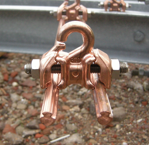 Closeup of a short, cut off section of the two contact wires of Dutch railway electrification. You can see the two notches in the wires where the mounting bits grab them and also the mounting bit which keeps the two wires neatly spaces. Everything is shiny copper (apart from the bolt and nuts that hold it all together).
