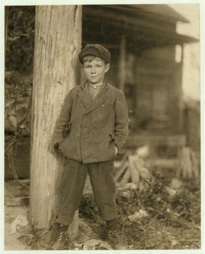  The image is a black and white photograph featuring a young boy standing in front of a wooden structure, leaning against it. He appears to be in the center of the frame, occupying a significant portion of the image. The boy is dressed in what seems to be early-20th century attire: he wears a long-sleeved shirt with rolled up sleeves and wide lapels, a dark tie, high-waisted trousers, and a dark cap.

The background reveals an older wooden building, possibly a house or store, with a porch extending across the front of the structure. A fence is visible in front of the building, suggesting a residential or semi-residential area. The boy's expression is neutral, and he stands with his legs crossed at the knee, hands hanging by his side.

There is no text present on the image to provide additional context. The style of the photograph, including the clothing and the monochromatic palette, suggests that it could be from the early 20th century. However, without specific visual clues or a time-stamp, an exact year for this photo cannot be determined. 