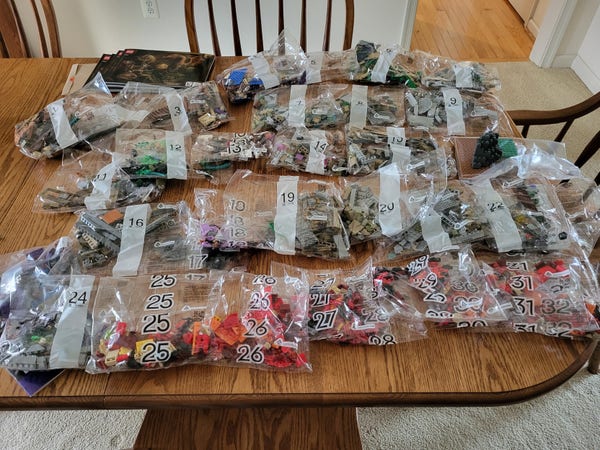 The Lego Dungeons & Dragons anniversary set, before construction. Plastic bags numbered 1 to 32 are laid out on a wooden table, each containing a variety of colorful plastic parts in shades of gray, red, green, and hints of blue and purple. 
