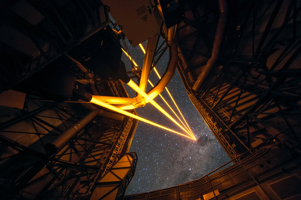 A photograph taken inside a telescope dome. The dome is partially open, and the telescope is firing four yellow laser beams that appear to converge somewhere on the starry sky.