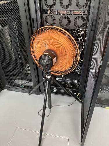 A large fan at the back of a server rack, blowing air into the rack in an attempt to cool a possibly overheating device