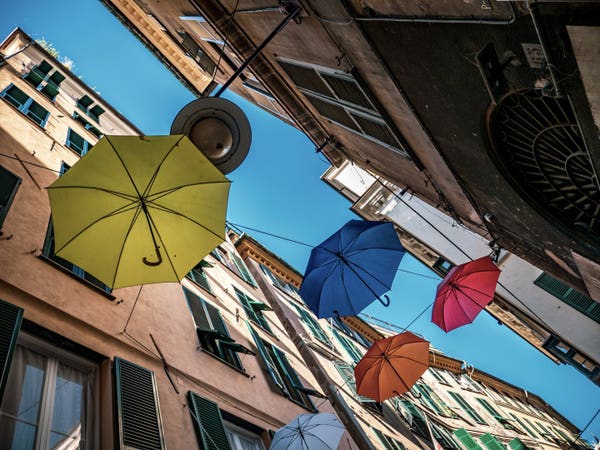 Photographed from a street looking up the houses left and right, with umbrellas in yellow, red, blue and orange hanging over the street. The sky above is blue