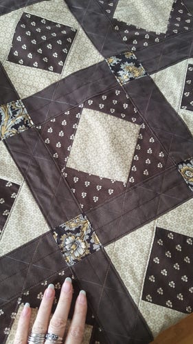 A photo of a quilt in progress. It is in shades of brown and cream, with historical prints of small flowers and leaves. There are both slightly drunk lines of stitching and fairly neat lines of chalk to denote where more stitching should be. There is a somewhat dense grid appearing, with lines going across and along strips of framing darker brown fabric, and criss-crossing lines going diagonally to those, catching the edges of what is called an economy block or square within a square. There is a determinedly grandmotherly legacy vibe to it!