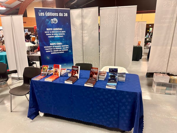 Un stand d'exposition de livres avec un panneau indiquant "Les Éditions du 38", présentant une variété de livres sur une table avec une nappe bleue. Des bannières promotionnelles se trouvent à l'arrière-plan.