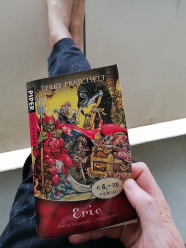 Die Taschenbuchausgabe des Buches "Eric" von Sir Terry Pratchett, der heute vor 76 Jahren geboren wurde.

Ich sitze auf dem Balkon und halte das Buch mit der rechten Hand in die Kamera, man sieht meine ausgestreckten Beine und einen Teil meiner nacktrn Füße. 