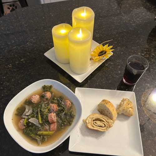 Kale soup with chicken sausage and Italian seasoning in a bowl with homemade herb garlic bread on a plate next to it. There’s also a glass of red wine and three candles and a sunflower on a plate. 