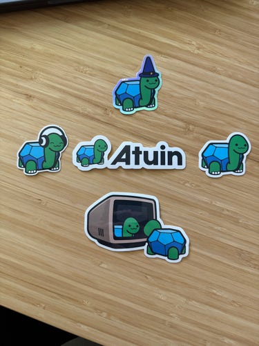 Five stickers from Atuin (Sync, search, and backup shell history with Atuin) on a wooden desk. All stickers have a cartoon turtle. One is wearing a wizard hat, another headphones, and one is looking at its reflection in a computer monitor. 