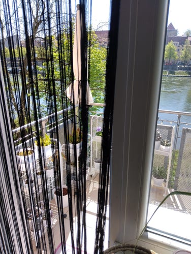 View from a window and open balcony glass door to a patio with a rail and potted plants. There is a string curtain separating inside from outside.