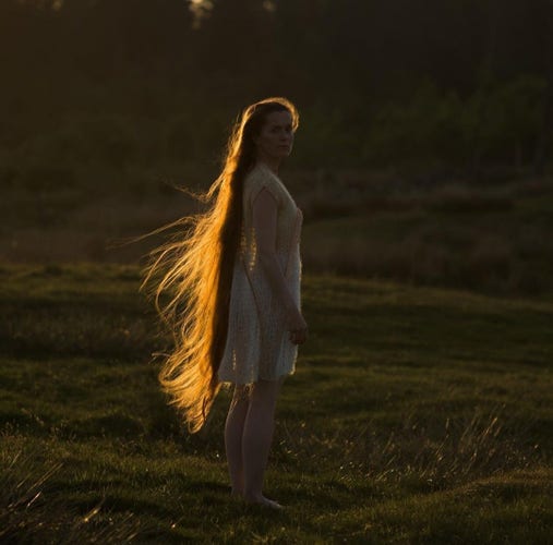 Photography. A color photo of a young woman in the sunlight. The scene is in semi-darkness. A young woman in a short, simple white summer dress is standing in a meadow. The little light falls on her beautiful, knee-length blonde hair from the side. She looks at us. A beautiful photo that plays with light and shadow and has a mystical, warm atmosphere.