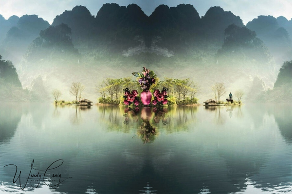 A serene landscape is mirrored perfectly on the surface of a tranquil lake, creating an almost symmetrical image featuring lush greenery and flowering trees. In the distance, limestone karst mountains emerge from the mist, enhancing the calm and mystical atmosphere of the scene.