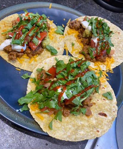 Three tacos with ground meat, shredded cheese, diced tomatoes, sour cream, hot sauce, and cilantro on a dark blue plate.