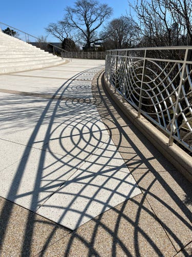 An outdoor staircase with a decorative metal railing casting intricate shadows on the ground. Trees without leaves can be seen in the background under a clear sky.