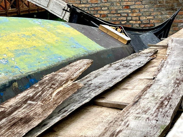 Colour photo of a gondola in bits, in the process of being repaired. Notable elements in the image are: the underside of a boat covered in blue, yellow and green paint; the pointed glossy black stern of another boat, which look like they should be attached to each other but are in fact not; a wooden ornament that looks like the shape of the top of Napoleon's hat; and three pieces of weathered salt-blemished driftwood. 