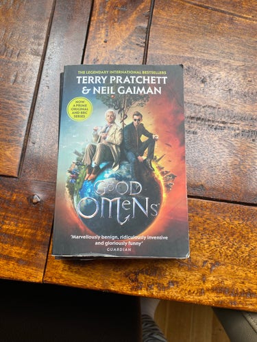 A paperback edition of "Good Omens" by Terry Pratchett & Neil Gaiman on a wooden surface.