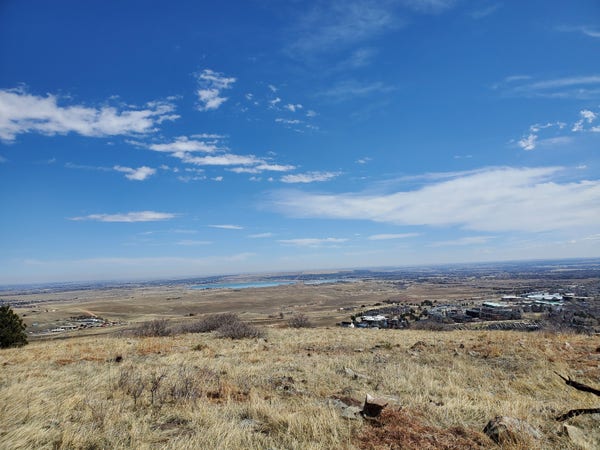 A panoramic view looking out across Boulder and over the plains. The sky is a beautiful blue studded with wispy clouds, some full and white, and some nearly transparent. The grass below is brown and crushed from recent winter snow. No snow can be seen nearby. Farr off in the distance a lake reflects the blue of the sky. In the middle ground on the right, the rooftops of houses can be seen. The ground drops away from the camera down a half mile slope before turning into rolling hills with a highway winding through.