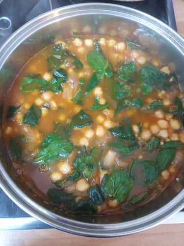 Photo of a saucepan cooking on the hob. Inside it is full of chickpeas, fresh spinach cooking in a tomato and broth sauce with onions, garlic, bay leaf, paprika sweet and spicy.
