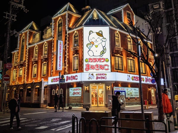Nighttime pic of a lot up corner 3 storey building with tower like portions and a huge waving cat poster/banner at the corner above the entrance glass doors to the karaoke place
Sannomiya Kobe