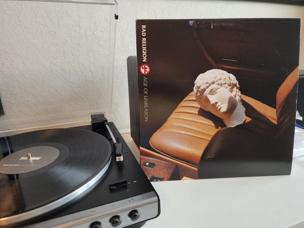Bad Religion - Age of Unreason album cover next to a record player. Cover is a statue head decapitated on the front seat of a car. Vinyl is black.