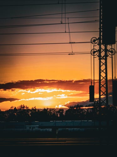 Subject:
The image captures a sunrise scene with the silhouette of power lines running across train tracks.

Image format: The image is in landscape format.

Genre: This falls under the silhouette photography genre, where the subjects are backlit and appear dark against a lighter background.

Background: The background features an orange and yellow sunset sky with scattered clouds, creating a dramatic effect.

Colors: The prominent colors are shades of orange, yellow, and black, creating a contrast between the silhouetted foreground and the bright, colorful sky.