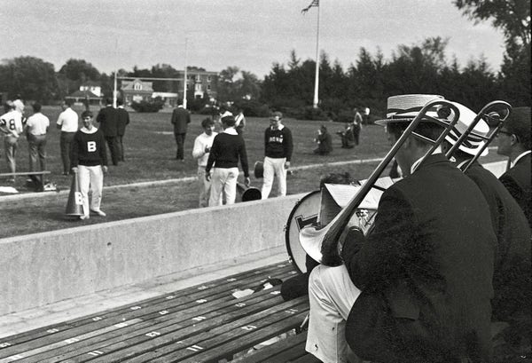Black and white photo of Brown Band trombone and bass drum players sitting at the bottom of the home field stadium bleachers, facing cheer leaders and coaches gathered near the north end of the field. The Band players wear dark blazers, white trousers, and straw hats with striped ribbon bands.