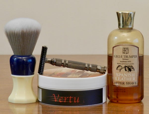A shaving bursh that has a grey synthetic knot with white tips and a hand with an extended white octagonal base with a round navy-blue top sections stands nexxtt to a tub of shaving soap shows black side label has "Vertu" printed in red. Lying on top is a bronze-colored double-open-comb safety razor. Next is a plastic bottle with a gold-cololred top and a label that reads "Geo. F. Trumper Spanish Leather After Shave."